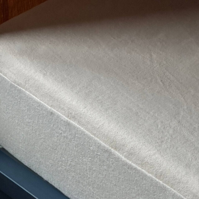 Eine Nahaufnahme eines ordentlich gemachten Bettes mit einem einfarbigen, hellen Spannbettlaken und einem Matratzenschoner nach Maß, der die Matratze bedeckt. Das Bettgestell ist dunkel, und ein Teil einer Holzwand trägt zur Erlebniswelt im Hintergrund bei.