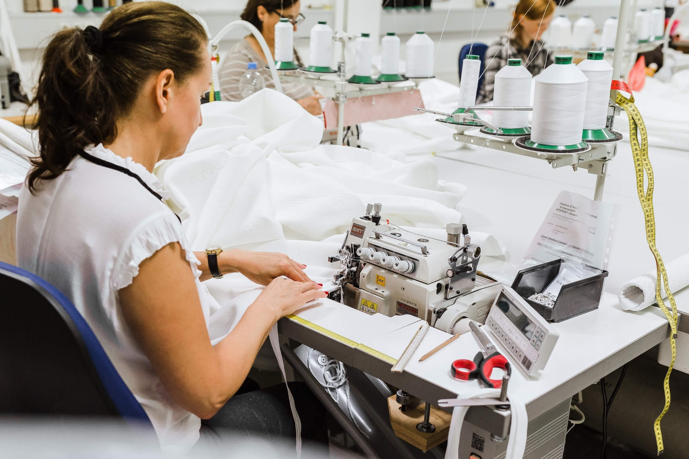 Eine Frau bedient eine Nähmaschine an einem Arbeitsplatz mit weißem Stoff, Garnspulen, Schere und Maßband; weitere Arbeiterinnen sind in der Textilwerkstatt der Erlebniswelt für Matratzenschoner nach Maß zu sehen.