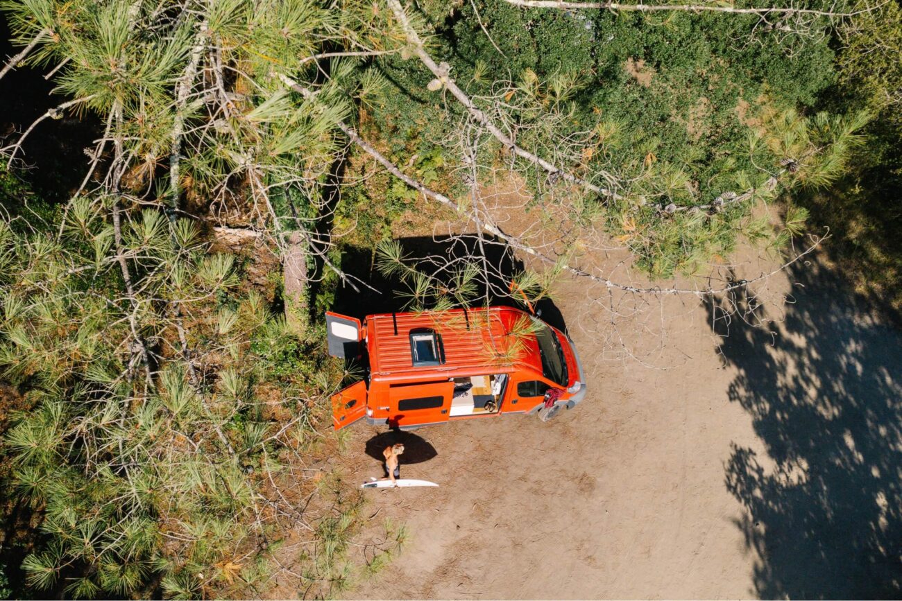 Luftaufnahme eines roten Lieferwagens, der auf einer unbefestigten Fläche neben grünen Bäumen geparkt ist. Eine Person steht draußen mit einem Surfbrett, das Sonnenlicht wirft Schatten. Die Türen des Lieferwagens sind offen, so dass eine Erlebniswelt im Freien entsteht, die erkundet werden kann.