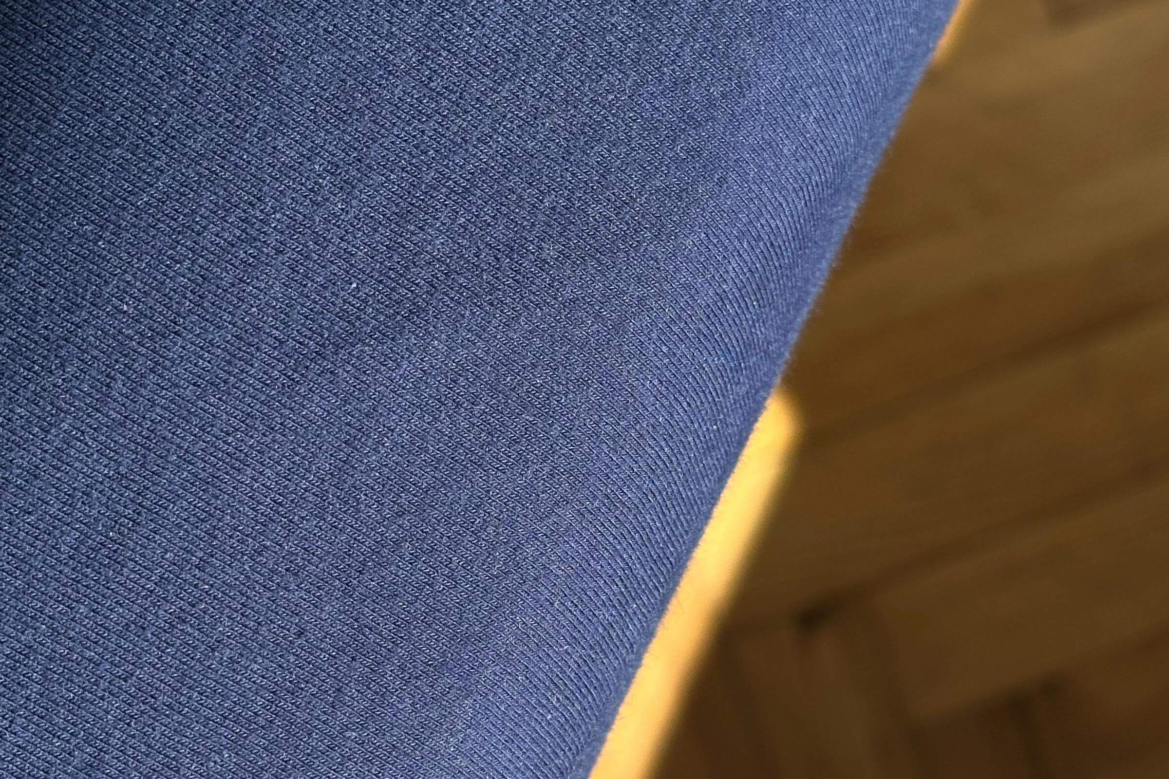 Nahaufnahme einer blauen Stoffoberfläche, möglicherweise Kleidung, mit einem Holzboden im Hintergrund. Das Bild ist schräg, und die Textur des Stoffes ist deutlich zu erkennen.