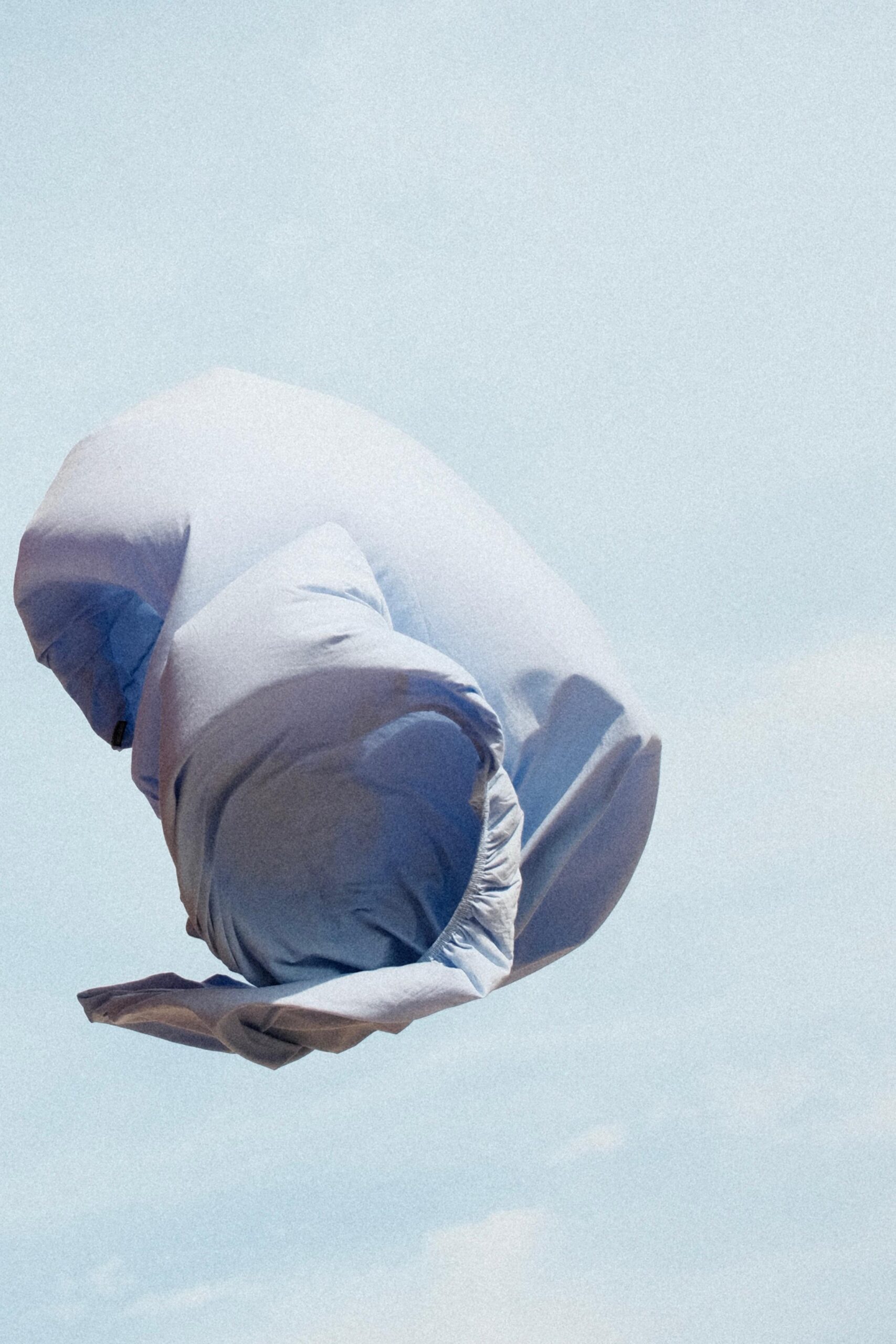 Ein hellblaues, zerknittertes Spannbettlaken nach Maß schwebt in der Luft vor einem blassblauen Himmel, so dass es aussieht, als würde es sich wellen oder vom Wind getragen werden.