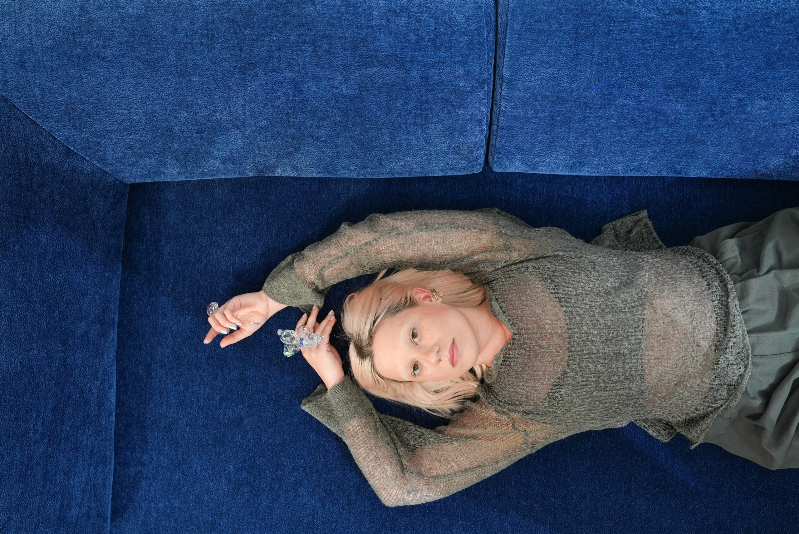 Eine Frau mit hellem Haar liegt auf einer blauen Couch und trägt ein durchsichtiges, langärmeliges Oberteil und einen Rock. Sie blickt nach oben, einen Arm über den Kopf gestreckt - die heitere Szene fängt den Look von AtelierEdition Hero ein.