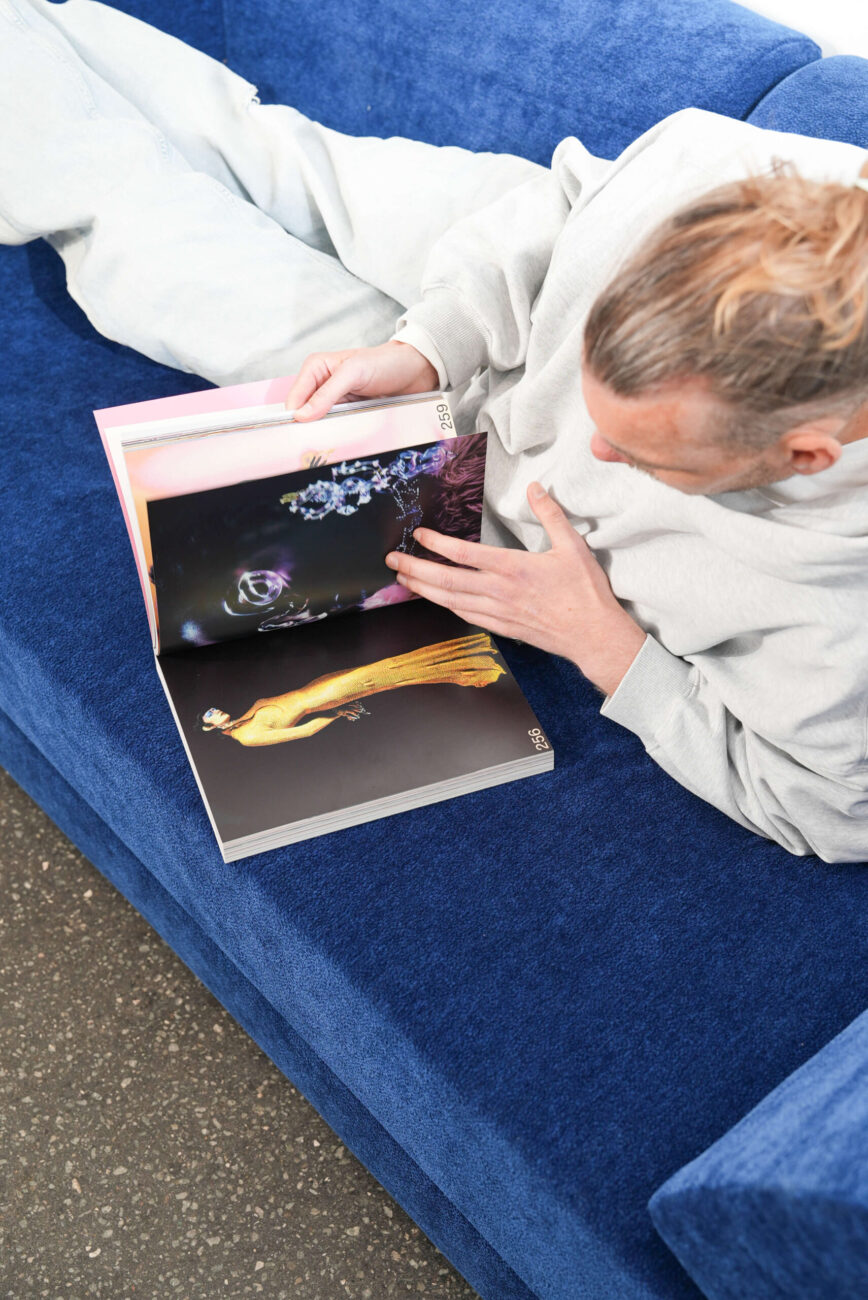 Eine Person mit hellbraunem Haar in einem Dutt liegt auf einer blauen Couch, trägt hellgraue Kleidung und blickt auf ein Modemagazin, das bunte Seiten mit einer Nahaufnahme und einem Modell in einem gelben Kleid zeigt.