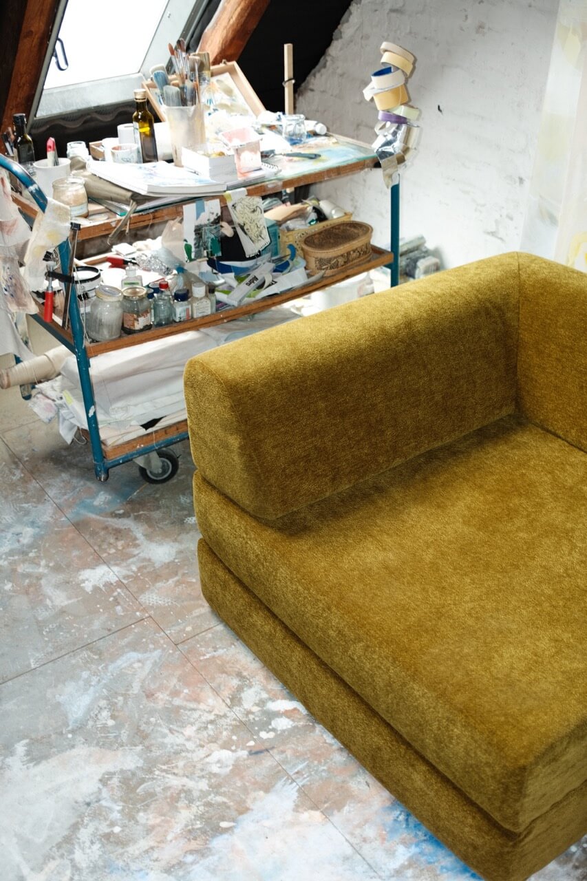 Ein senfgelber Sessel steht auf einem mit Farbe bespritzten Boden in der Nähe eines Fensters. Dahinter steht ein Metallwagen mit Malutensilien, Pinseln, Flaschen und Gläsern, die an ein Künstleratelier erinnern.