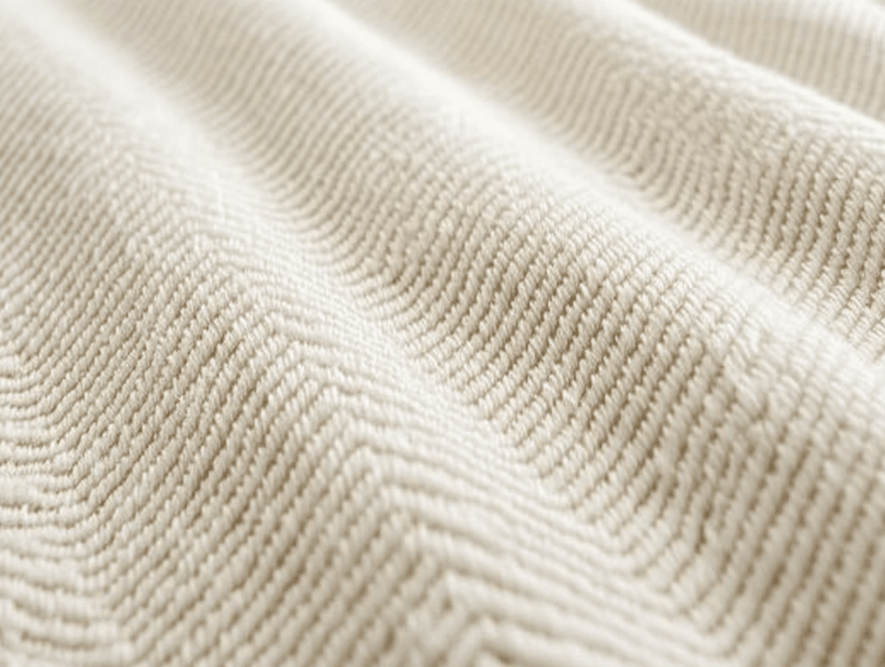 Nahaufnahme eines cremefarbenen, strukturierten Stoffes, der ein subtiles Zickzack- oder Chevron-Muster aufweist, mit Köperbindung, die sanfte, wellenförmige Rillen und weiche Schatten auf der Oberfläche erzeugt.