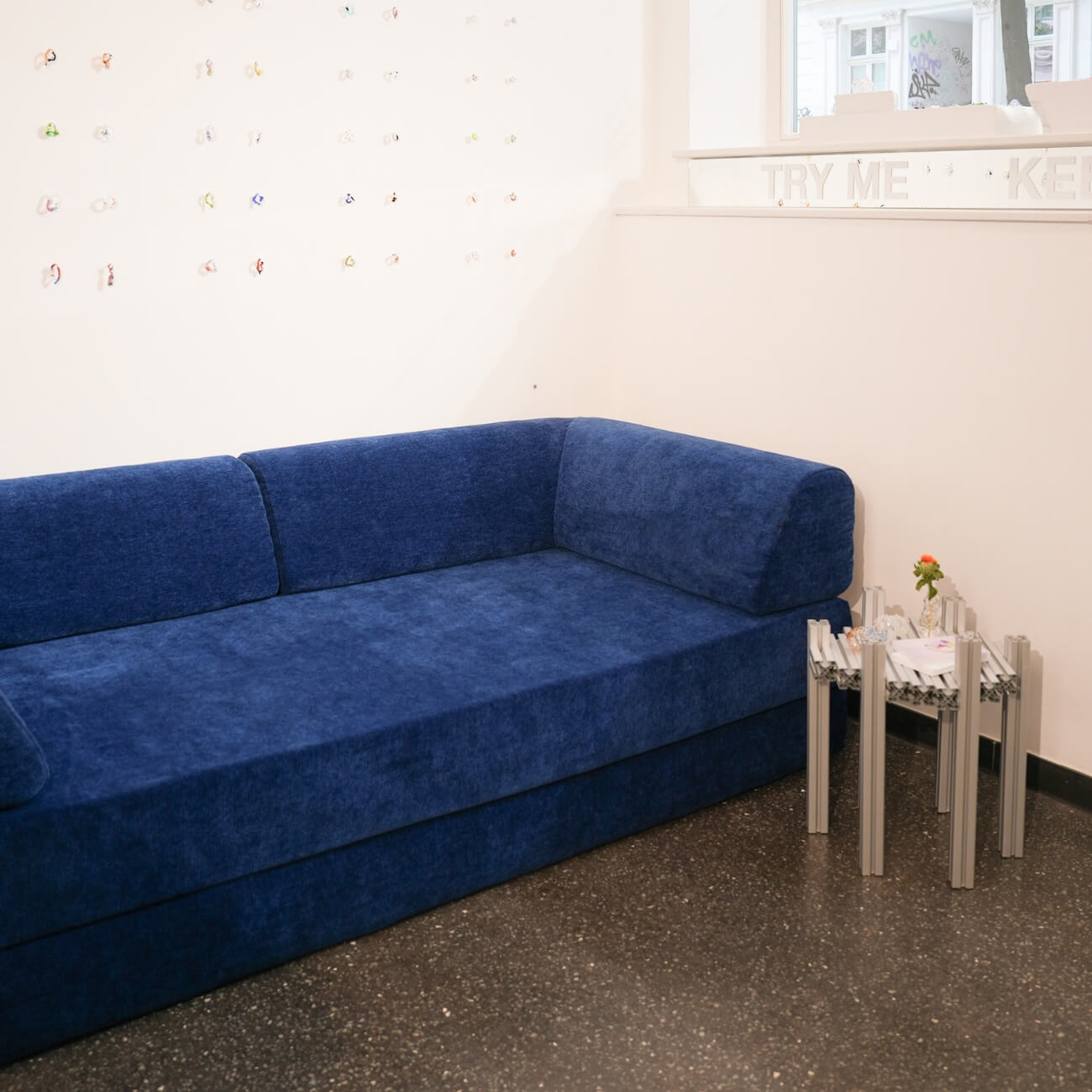 Ein modernes Zimmer mit einem blauen Samtsofa, einem kleinen Beistelltisch mit Zeitschriften und einer einblütigen Blume in einer Vase und einer weißen Wand mit kleinen Dekorationsgegenständen. Licht fällt durch ein Fenster über dem Sofa ein.