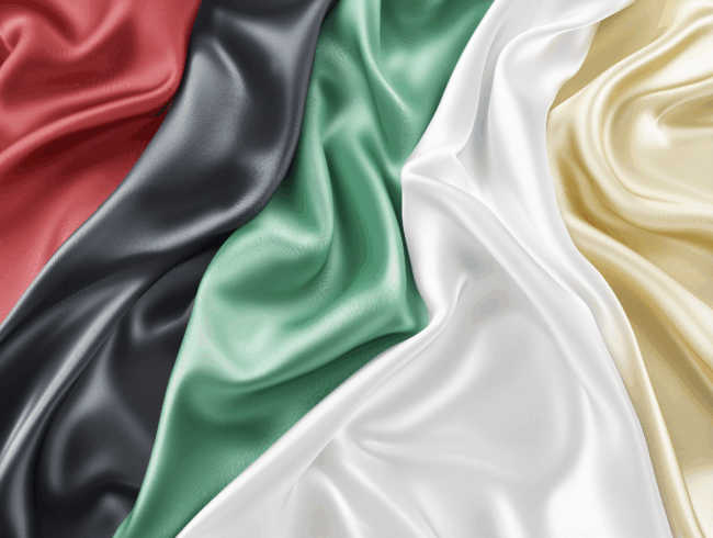 Nahaufnahme eines fließenden Acetat-Stoffes mit fünf verschiedenen Farben: rot, schwarz, grün, weiß und gelb, angeordnet in diagonalen Streifen. Die seidenartige Textur erzeugt weiche Falten und Highlights.