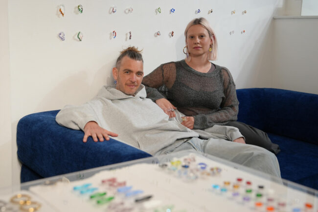 Zwei Personen sitzen auf einer blauen Couch, die eine in einem grauen Kapuzenpulli, die andere in einem dunklen Netzoberteil, zurückgelehnt. Im Vordergrund sind bunte Schmuckstücke ausgestellt und an der weißen Wand hinter ihnen angebracht.