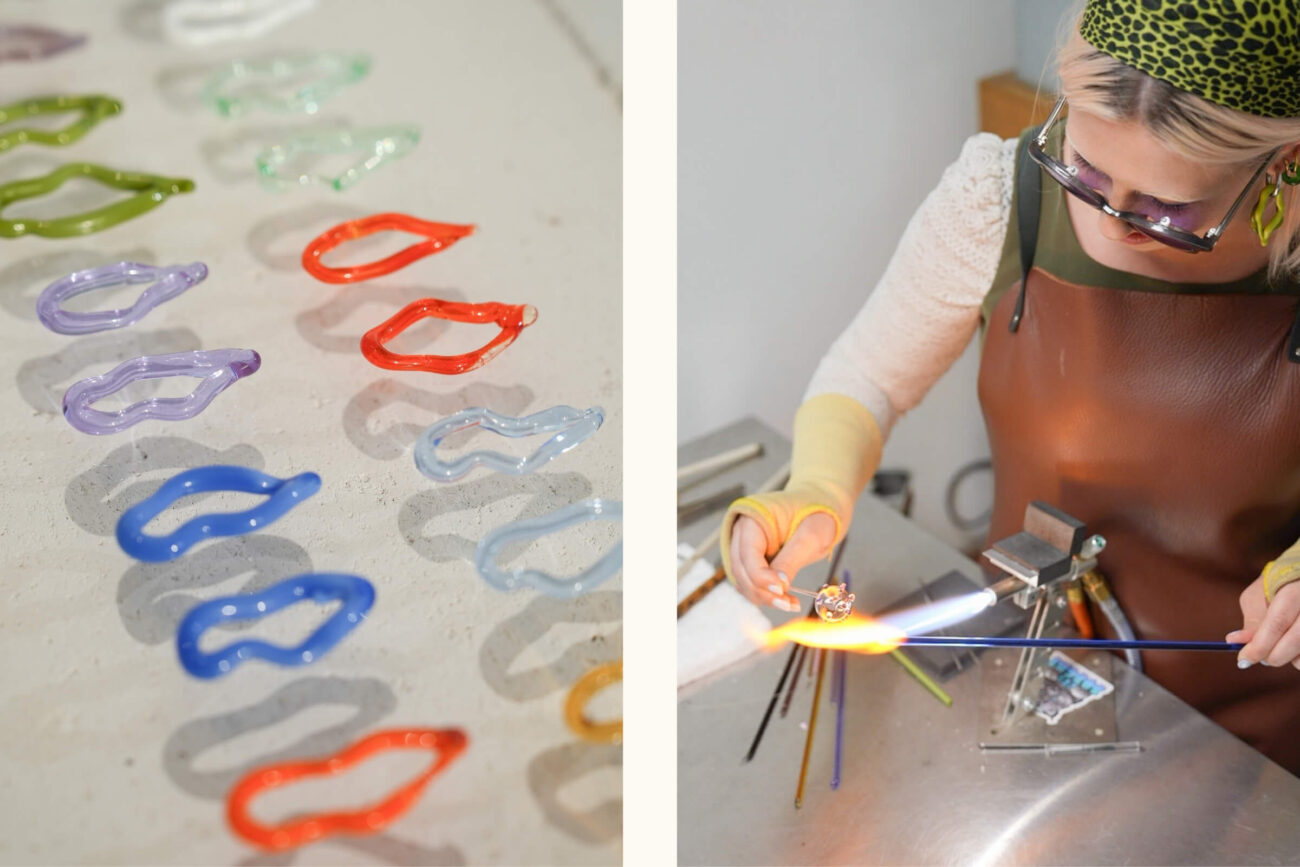 Auf der linken Seite sind bunte Glasformen, die an Lippen erinnern, auf einer Fläche angeordnet. Rechts formt eine Person, die eine Brille und eine Schürze trägt, an einer Werkbank Glas mit einem Brenner.