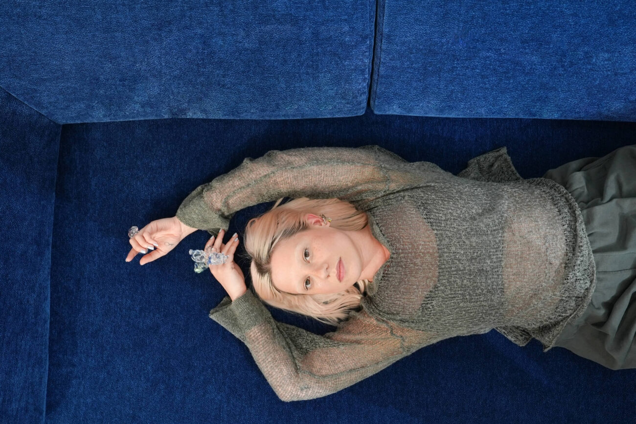 Eine Frau mit hellem Haar liegt auf einem dunkelblauen Sofa, trägt ein durchsichtiges graues Oberteil und hält eine Hand über ihren Kopf, während sie nach oben blickt.