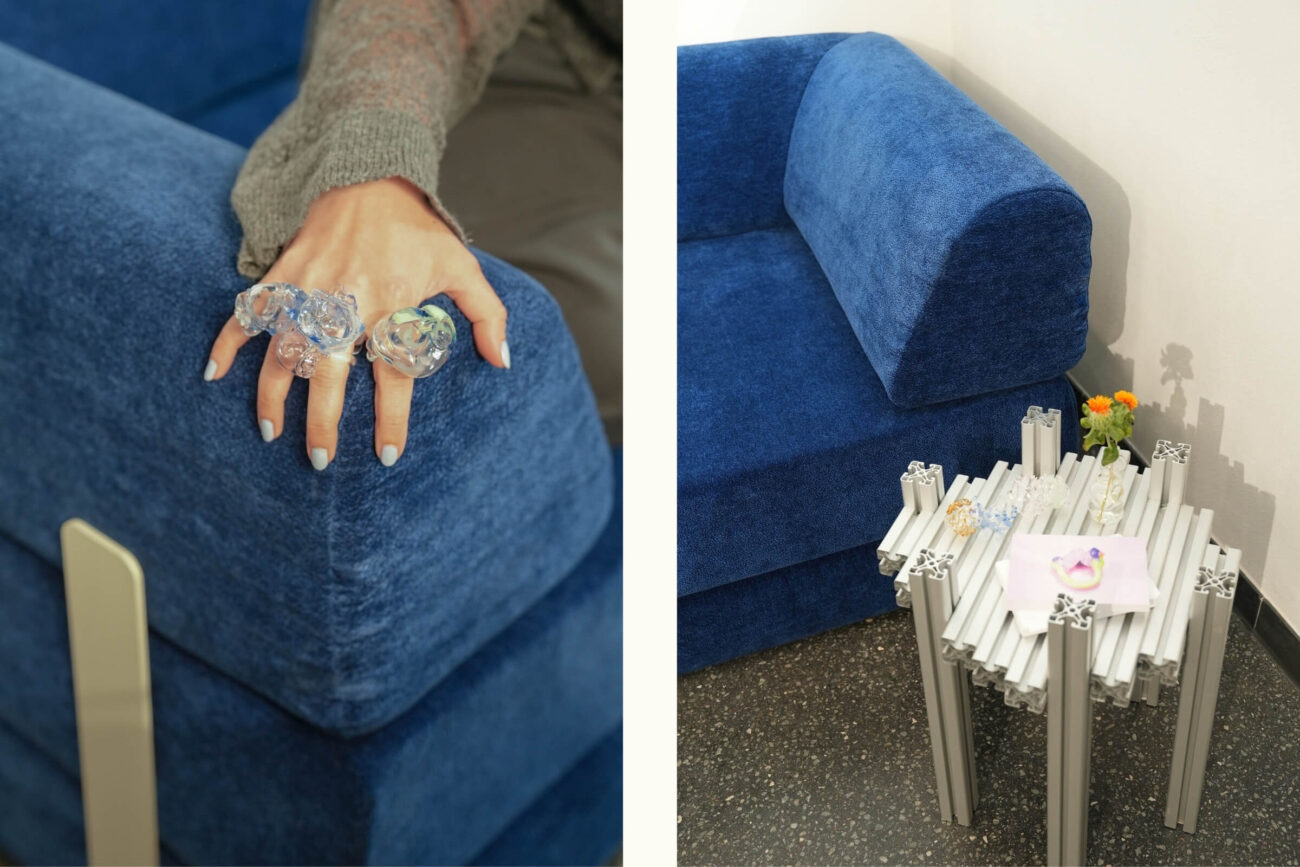Links: Eine Hand mit großen, klaren, skulpturalen Ringen ruht auf einer blau gepolsterten Couch. Rechts: Eine moderne blaue Couch neben einem kleinen silbernen Tisch im Industriestil, auf dem Ringe und eine kleine Vase mit Blumen stehen.