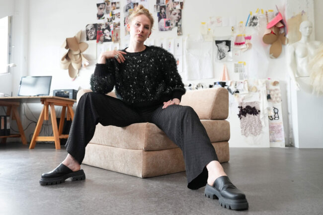 Eine Frau mit kurzen blonden Haaren sitzt selbstbewusst auf einem beigen Hocker in einem Kreativstudio. Sie trägt einen schwarzen strukturierten Pullover, eine schwarze Hose und klobige schwarze Schuhe. Kunstwerke und Stoffe schmücken die Wand hinter ihr.