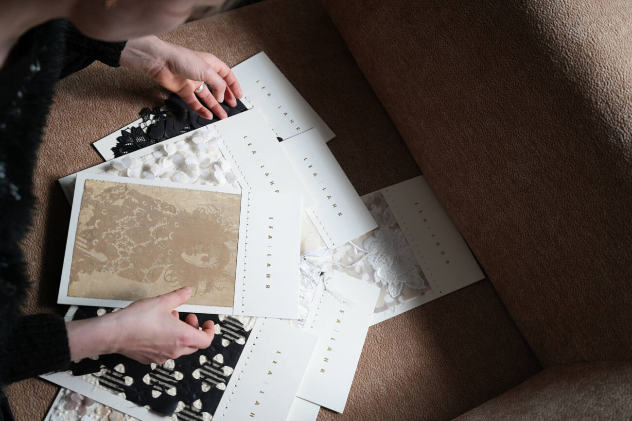Eine Person, die auf einer Couch sitzt, ordnet mehrere strukturierte und gemusterte Stoffmuster auf weißen Karten mit der Aufschrift SELANAH an. Zu den Mustern gehören schwarze, weiße und beigefarbene Designs.