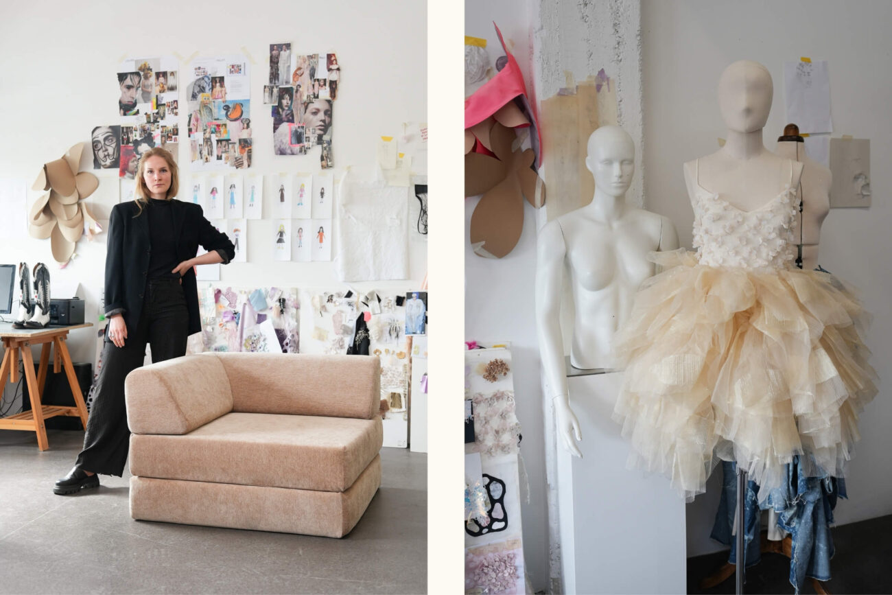 Eine Frau in schwarzer Kleidung steht an einem beigen Stuhl in einem Atelier mit Modeskizzen an der Wand. Neben ihr zeigen zwei Kleiderformen ein weißes Tüllkleid und ein Mieder inmitten von Stoff- und Musterteilen.