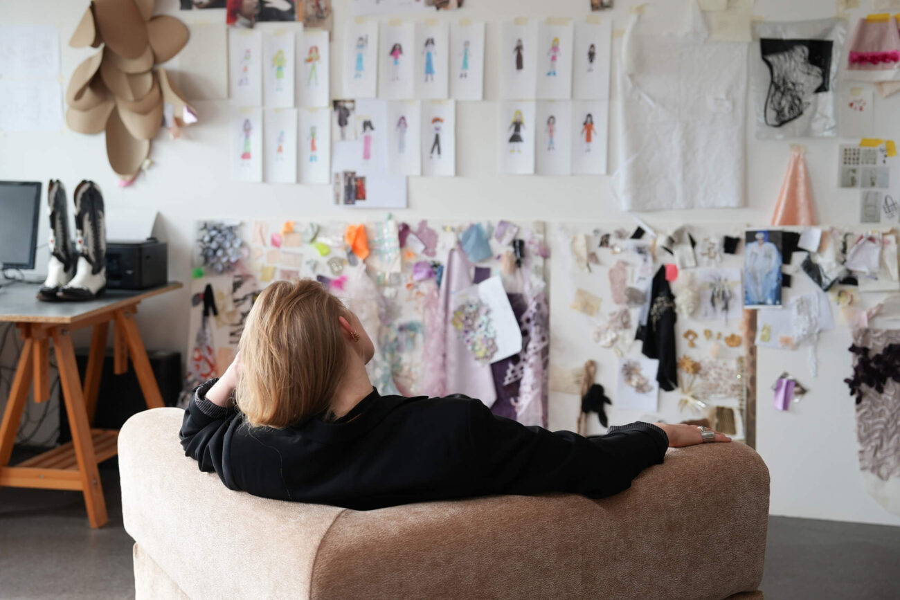 Eine Person mit blondem Haar sitzt auf einer beigen Couch und blickt auf eine Wand, die mit Modeskizzen, Stoffmustern und Designmaterialien in einer kreativen Atelierumgebung bedeckt ist.