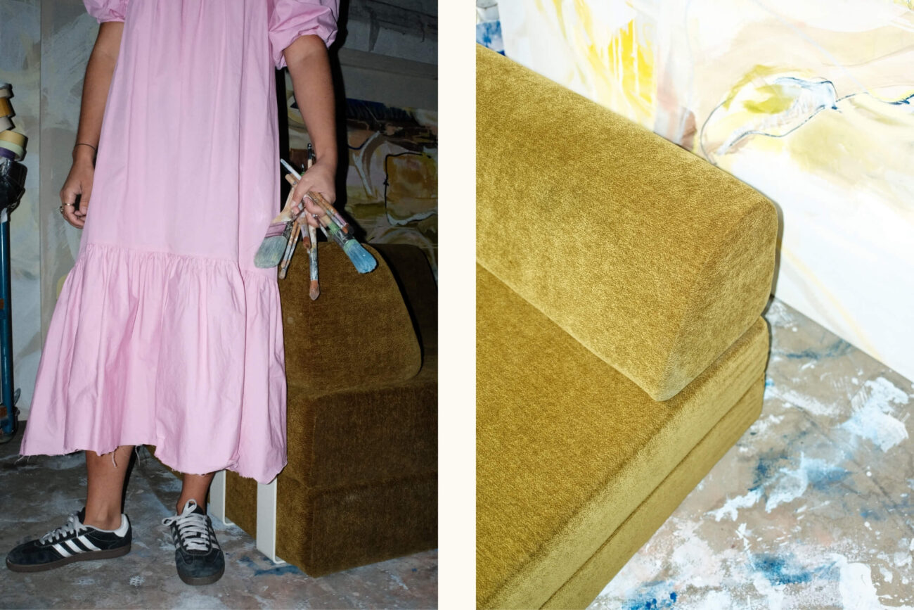 Eine Person in einem rosa Kleid und schwarzen Turnschuhen hält Pinsel neben einer senfgelben Couch. Der Boden ist mit Farbe bespritzt. Eine Nahaufnahme der Couch erscheint auf der rechten Seite des Bildes.