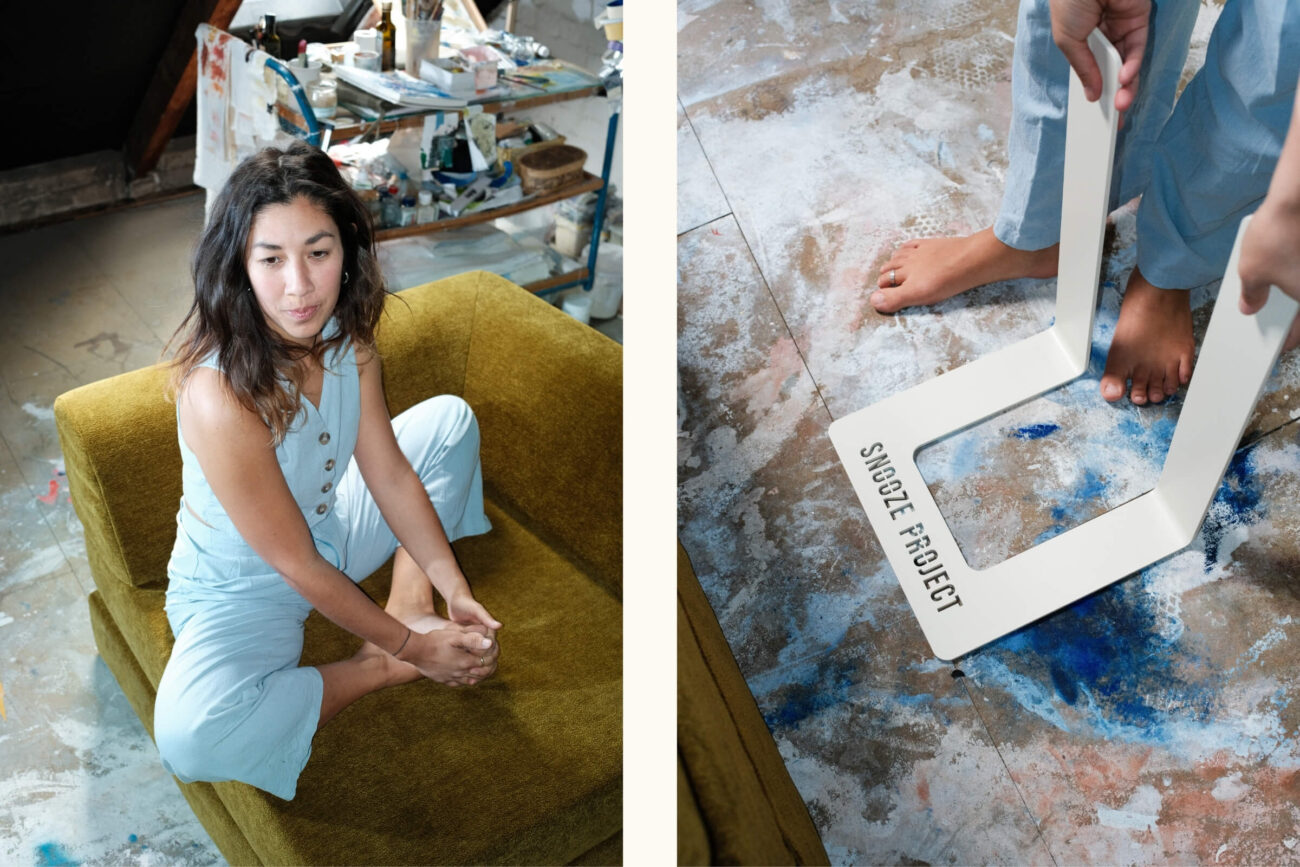 Eine Frau in einem hellblauen Overall sitzt nachdenklich auf einem senfgelben Stuhl in einem Kunstatelier. Neben ihr sieht man in einer Nahaufnahme nackte Füße, die in einem weißen Rahmen mit der Aufschrift "Snooze Project" auf einem mit Farbe bespritzten Boden stehen.