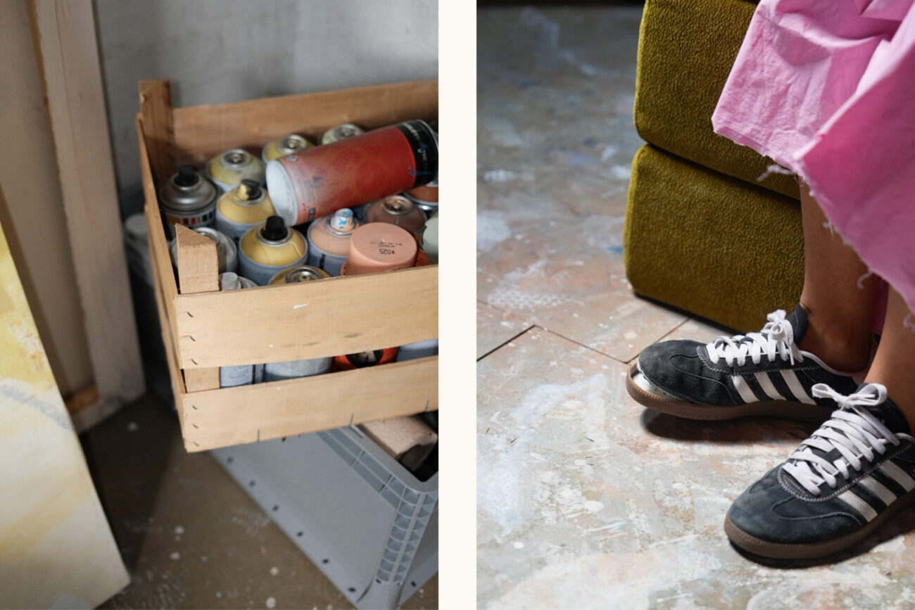 Auf dem Boden steht eine Holzkiste mit Farbsprühdosen; daneben steht eine Person in schwarzen Adidas-Turnschuhen und einem rosa Rock auf einer mit Farbe bespritzten Fläche neben einem grünen Stuhl.
