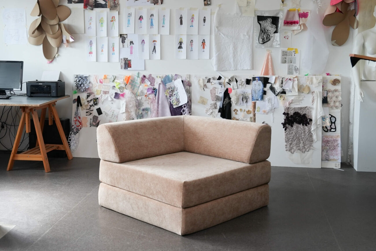 Ein moderner, beigefarbener Polsterstuhl steht in einem Atelier mit einer Wand, die mit Modeskizzen, Stoffmustern und Designnotizen bedeckt ist. Auf der linken Seite ist ein hölzerner Schreibtisch mit einem Drucker zu sehen.