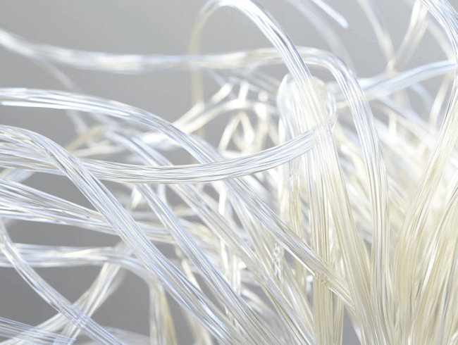 Nahaufnahme mehrerer durchsichtiger, flexibler Chemiefasern oder Glasfasern, die vor einem weichen, hellen Hintergrund ineinander verschlungen sind und ein abstraktes und kompliziertes Muster bilden.