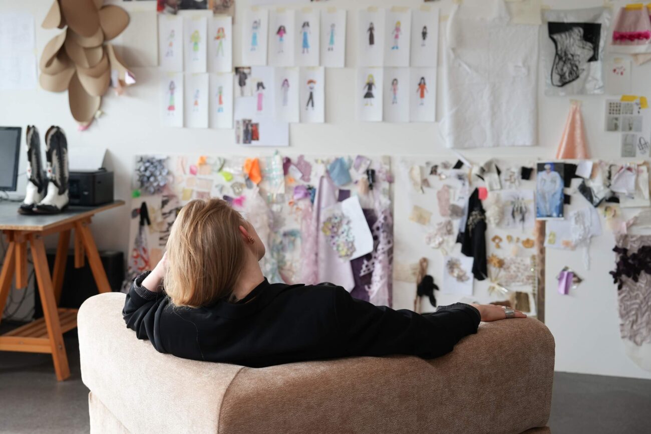 Eine Person mit hellem Haar sitzt auf einem beigen Sofa und blickt auf eine Wand mit Modeskizzen, Stoffmustern und Inspirationstafeln in einem Kreativstudio.