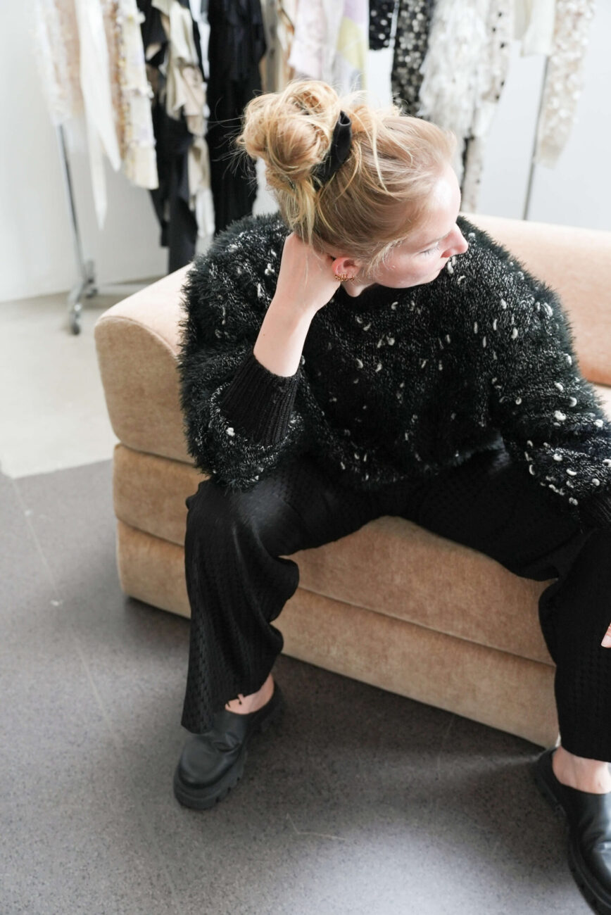 Eine Frau mit blondem Dutt, schwarzem Pullover und schwarzer Hose sitzt auf einer beigen Couch und blickt zur Seite, wobei sie ihre Hand in den Nacken legt. Im Hintergrund hängen Kleidungsstücke auf Ständern.