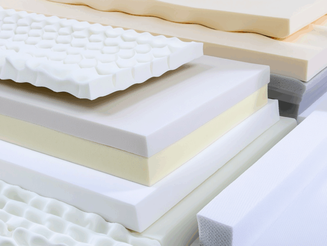 Stapel verschiedener Komfortschaum-Matratzenschichten in unterschiedlichen Dicken, Farben und Strukturen, einschließlich glatter, gewellter und wabenförmiger Oberflächen, in einem geordneten Stapel in Nahaufnahme angeordnet.