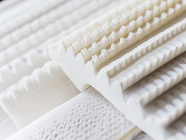 Nahaufnahme mehrerer weißer Schaumstoffmatratzenauflagen oder -polster, einschließlich Talalay-Latex-Varianten, mit unterschiedlichen Strukturmustern wie Rillen und Unebenheiten, nebeneinander angeordnet.