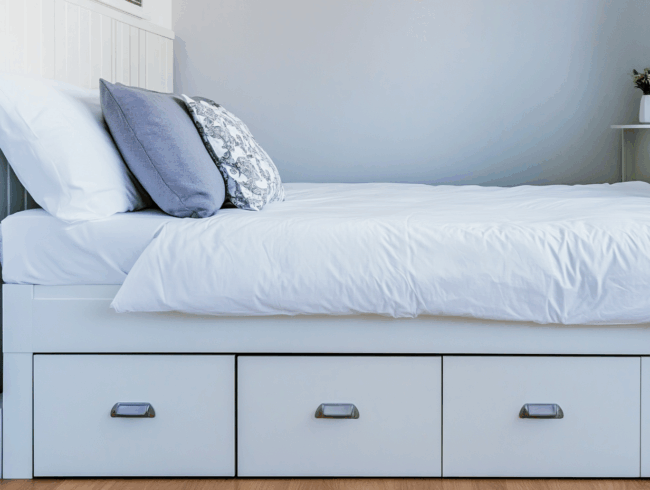 Ein ordentlich gemachtes Bett mit weißem Bettzeug und grauen Kissen steht auf einem weißen Funktionsbett mit drei geschlossenen Schubladen in einem hellen, minimalistischen Schlafzimmer mit hellen Wänden und Holzfußboden.