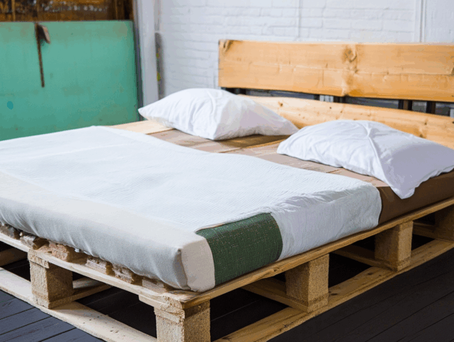 Ein Palettenbett mit einem Holzpalettenrahmen, einer Matratze, weißen Laken und zwei weißen Kissen steht vor einer weißen Backsteinwand in einem minimalistischen Raum.