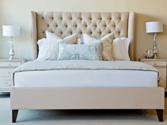 Ein ordentlich gemachtes Polsterbett mit einem getufteten beigen Kopfteil, Zierkissen in Weiß und Hellblau, flankiert von zwei weißen Nachttischen mit silbernen Lampen, alles in einem sanft beleuchteten, eleganten Schlafzimmer.