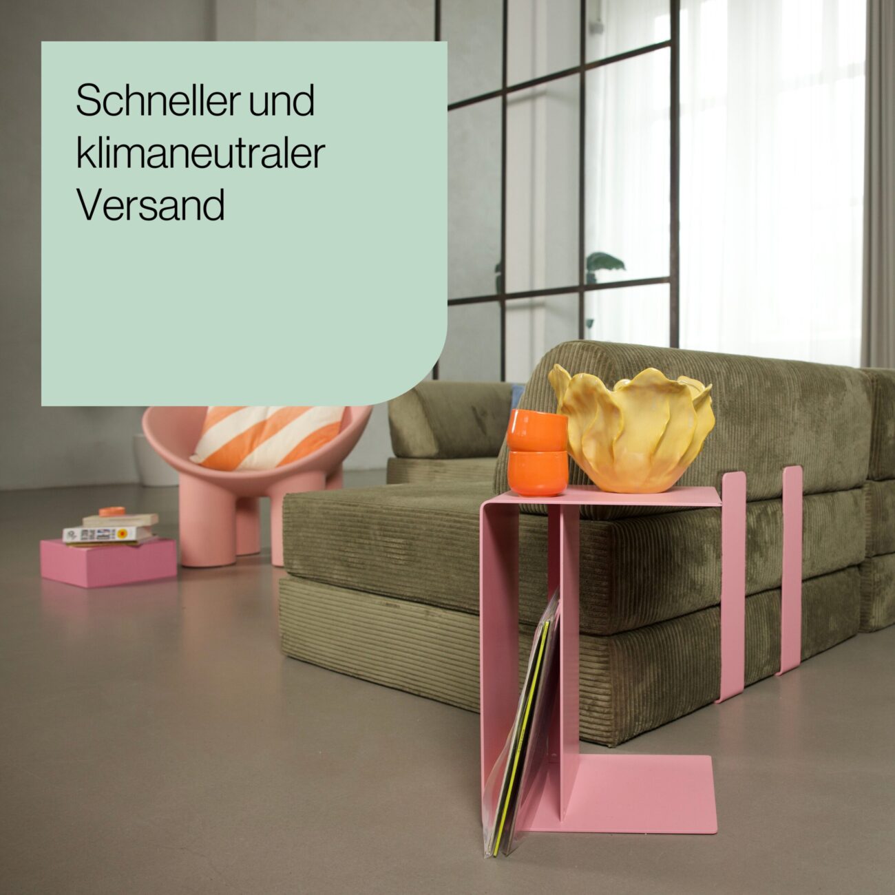 Tade_Produkt Mood_03 Ein modernes Wohnzimmer mit einem grünen Sofa, einem rosa Beistelltisch mit einer orangefarbenen Tasse und einer gelben Vase sowie einem rosa Stuhl im Hintergrund. Der Text lautet: Schneller und klimaneutraler Versand.