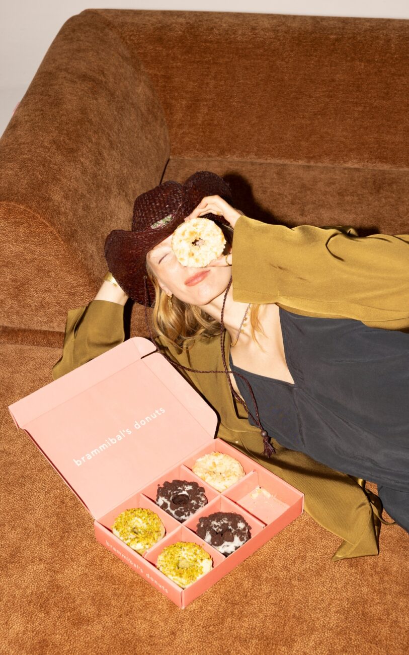 Eine Frau mit einem braunen Hut und einer grünen Jacke sitzt auf einer braunen Couch und hält sich eine Leckerei von Brammibal's Donuts vor das Gesicht. Auf der Couch neben ihr steht eine geöffnete rosa Schachtel mit verschiedenen Donuts vom Snooze Project.