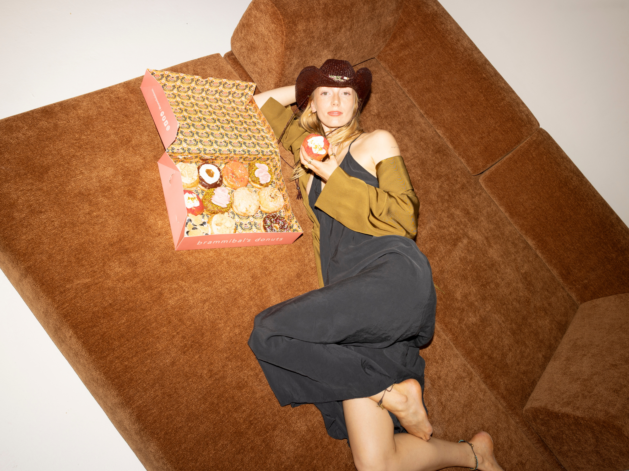 Eine Frau in einem schwarzen Kleid und einem braunen Cowboyhut lümmelt auf einer braunen Couch von Snooze Project und hält einen Donut in der Hand. Neben ihr steht eine offene rosa Schachtel mit verschiedenen Brammibal's-Donuts. Die Szene ist hell beleuchtet.