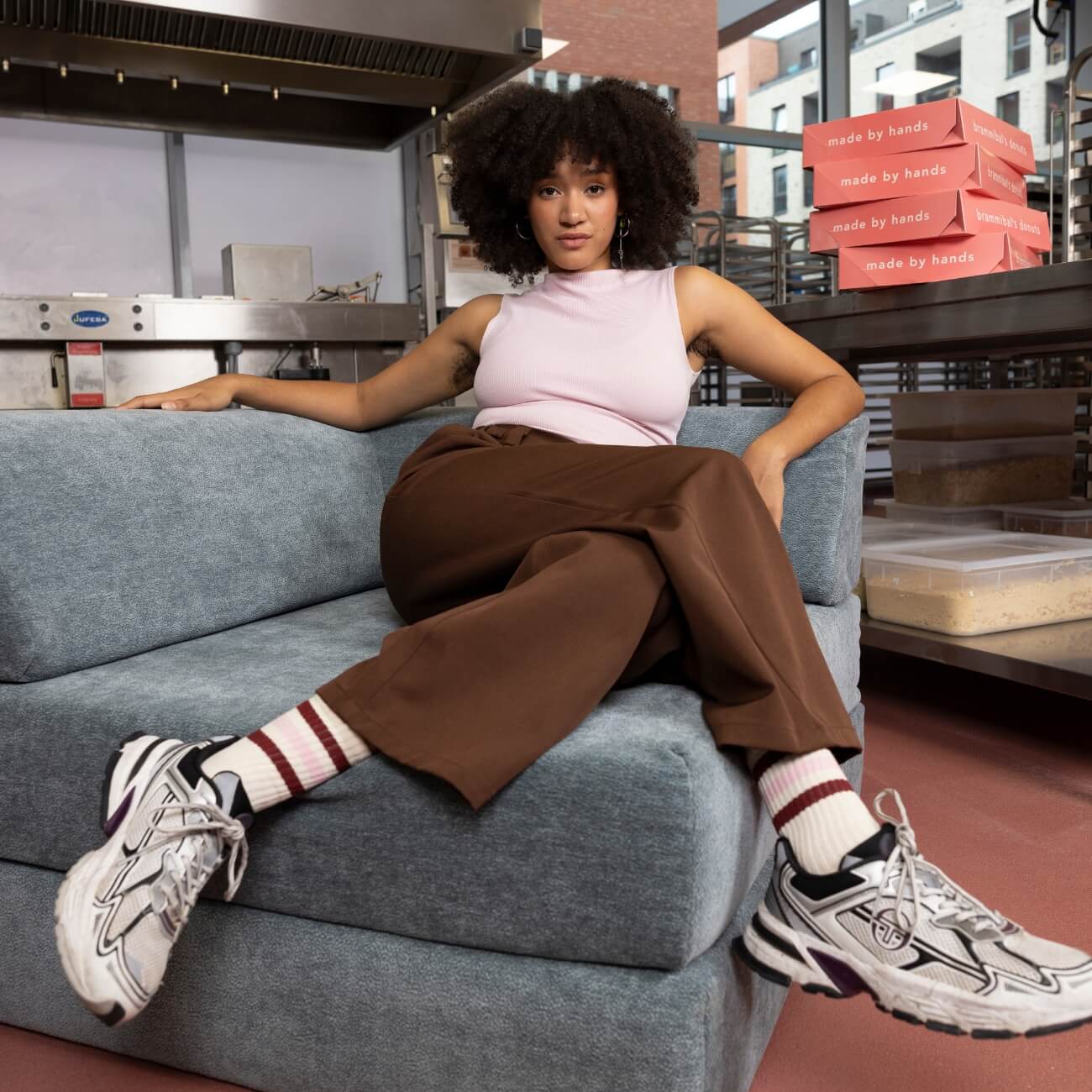 Eine Frau mit lockigem Haar sitzt auf einem grauen Sofa in einer modernen Küche und trägt ein ärmelloses rosa Oberteil, eine braune Hose, gestreifte Socken und Turnschuhe. Im Hintergrund stapeln sich rosa Pizzakartons mit der Aufschrift "von Hand gemacht".