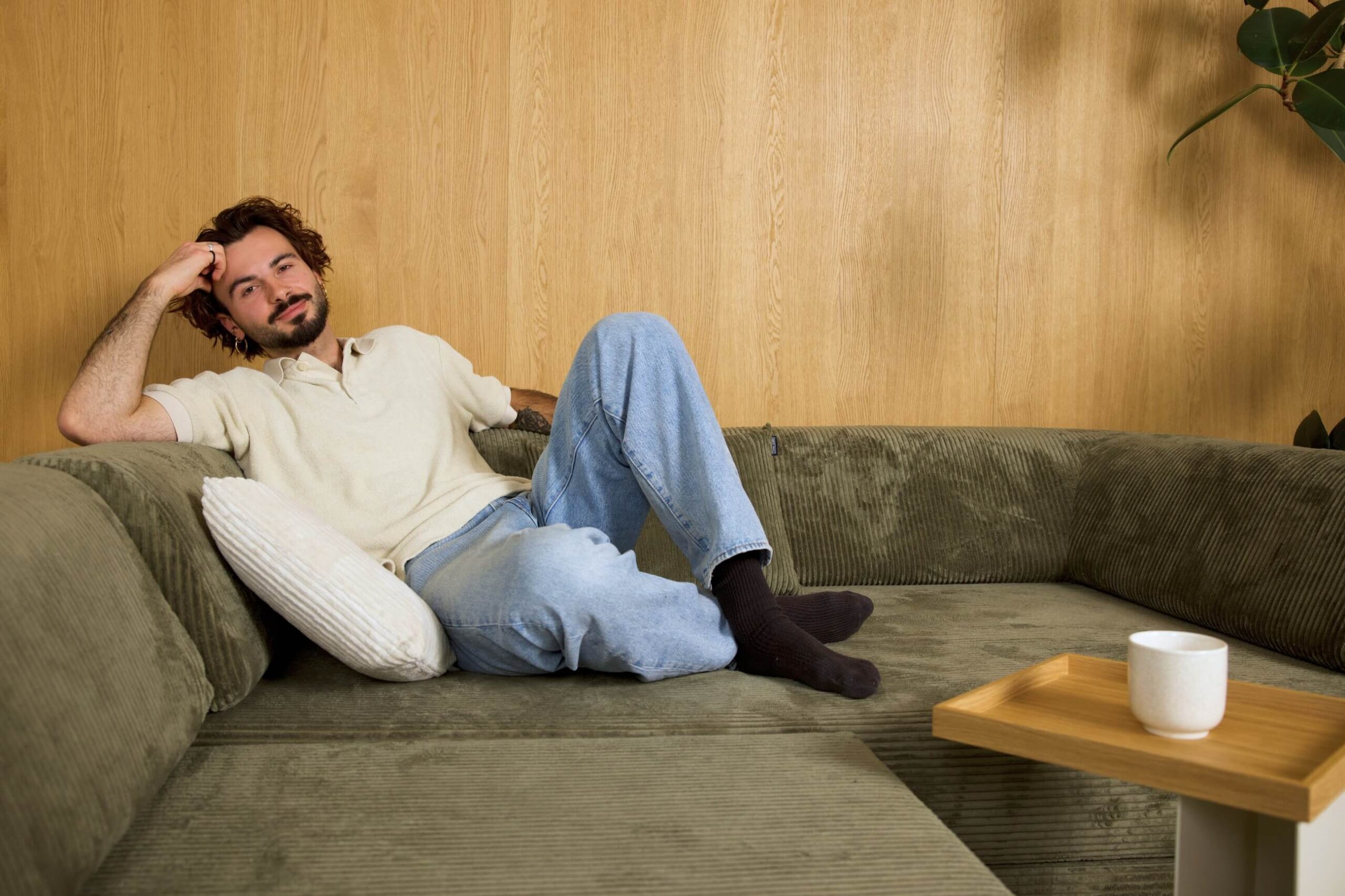 Ein Mann mit lockigem Haar und Bart sitzt auf einem grünen Sofa, das mit einem weißen Kissen abgestützt ist. Er trägt ein helles Hemd, blaue Jeans und schwarze Socken. Ein Kaffeebecher steht auf einem kleinen Holztisch in der Nähe.