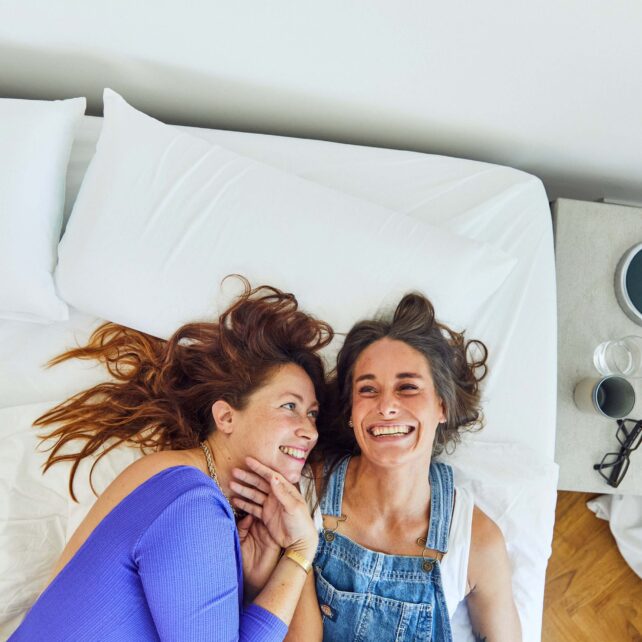 Zwei Frauen liegen auf einem Bett, lächeln sich an und schauen sich an. Die eine trägt ein blaues Oberteil, die andere einen Jeans-Overall. Sie wirken glücklich und entspannt, ihre Köpfe liegen eng beieinander auf dem weißen Bettzeug.