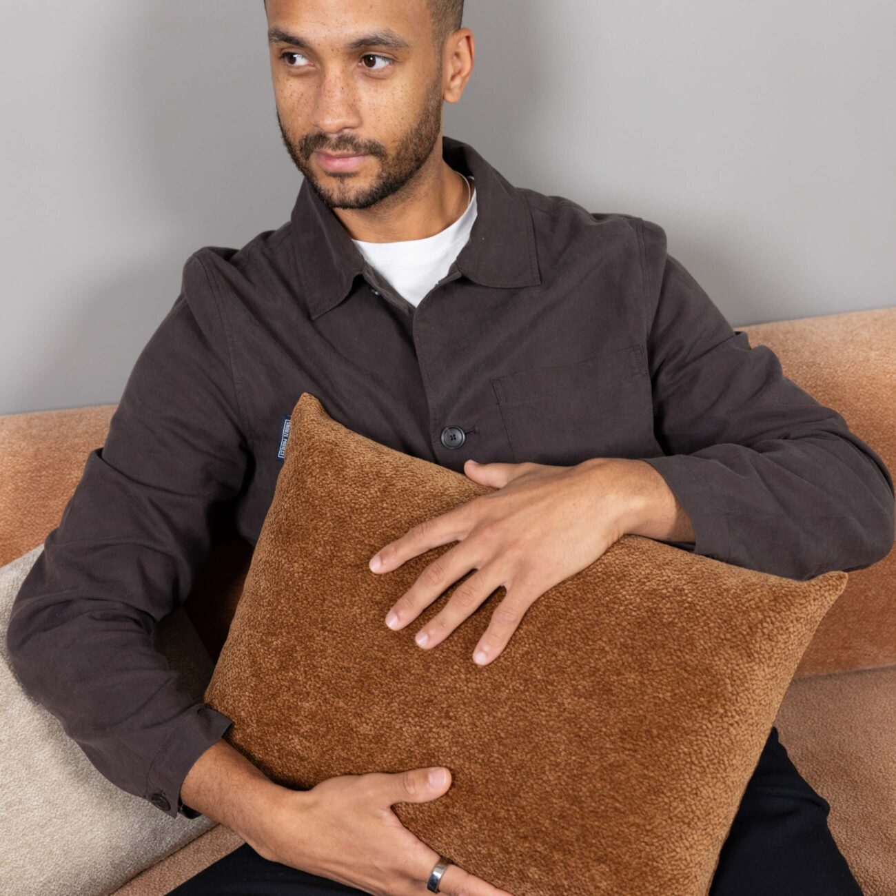 Ein Mann sitzt auf einem beigen Sofa, trägt eine dunkle Jacke über einem weißen Hemd und hält ein braunes, strukturiertes Kissen in seinen Armen, während er leicht zur Seite schaut.