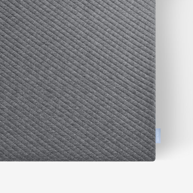 Nahaufnahme einer rechteckigen dunkelgrauen Matte mit diagonalen, strukturierten Linien auf weißem Hintergrund. Ein kleines hellblaues Stoffschildchen ist am Rand in der rechten unteren Ecke angebracht.