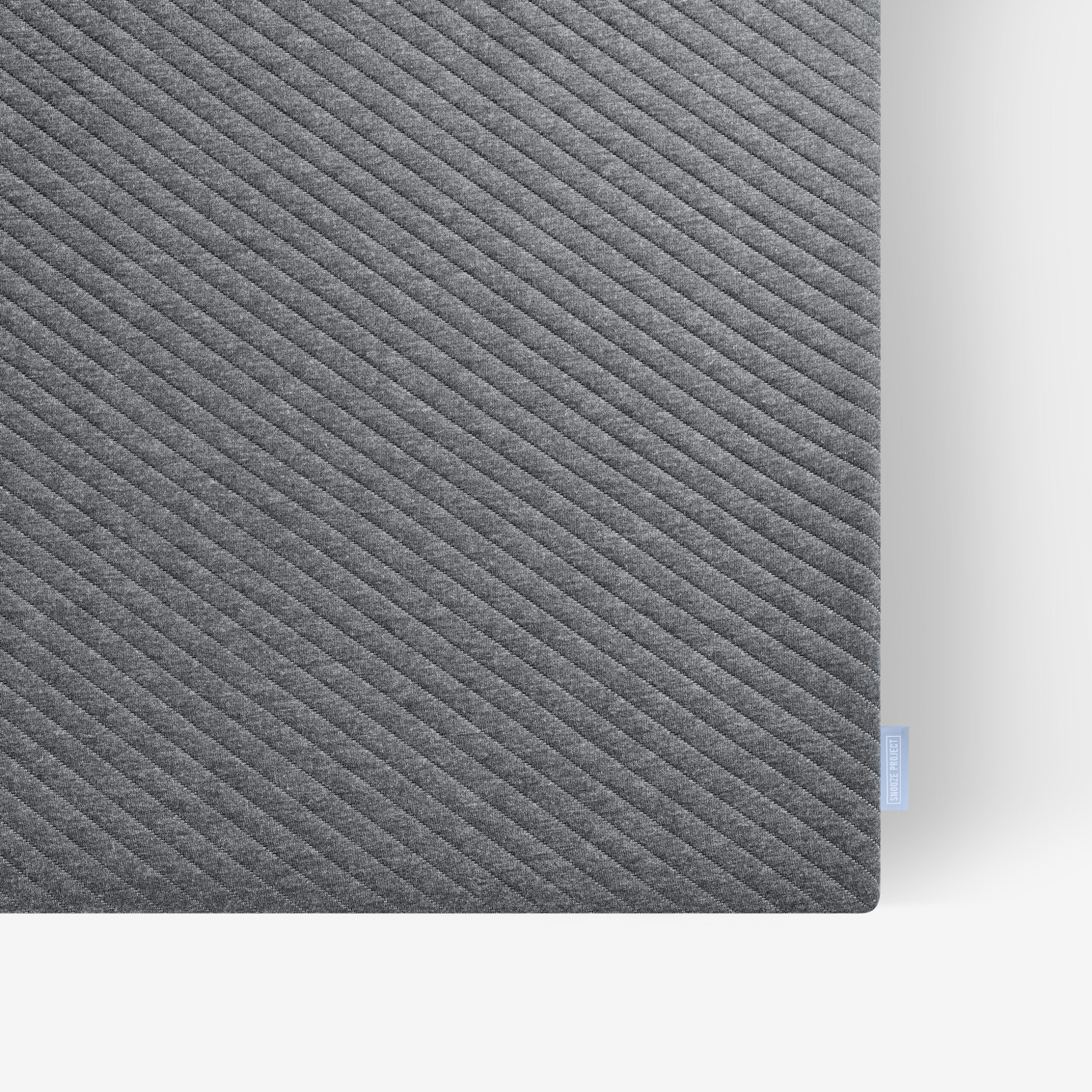 Nahaufnahme einer rechteckigen dunkelgrauen Matte mit diagonalen, strukturierten Linien auf weißem Hintergrund. Ein kleines hellblaues Stoffschildchen ist am Rand in der rechten unteren Ecke angebracht.