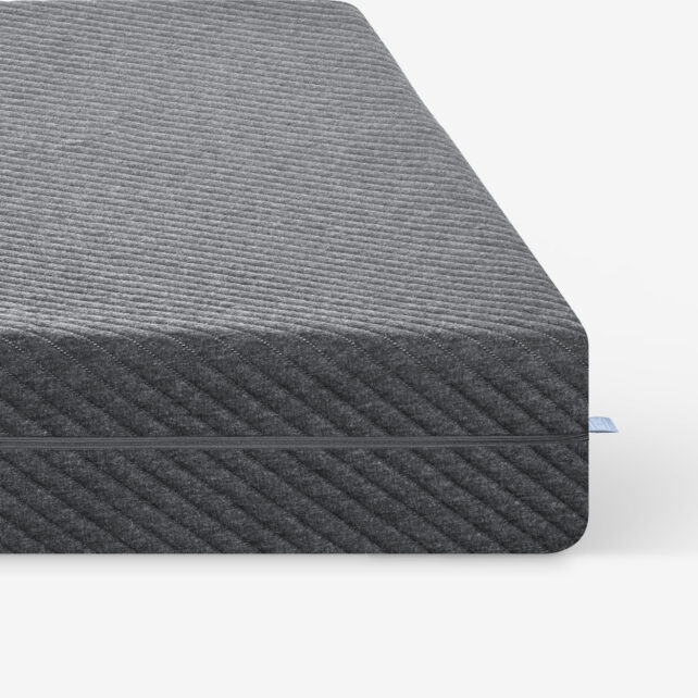 Nahaufnahme der Ecke einer dunkelgrauen, strukturierten Matratze mit einem dezenten diagonalen Streifenmuster vor einem schlichten weißen Hintergrund. Ein kleines blaues Stoffetikett ist an der Matratzennaht sichtbar.