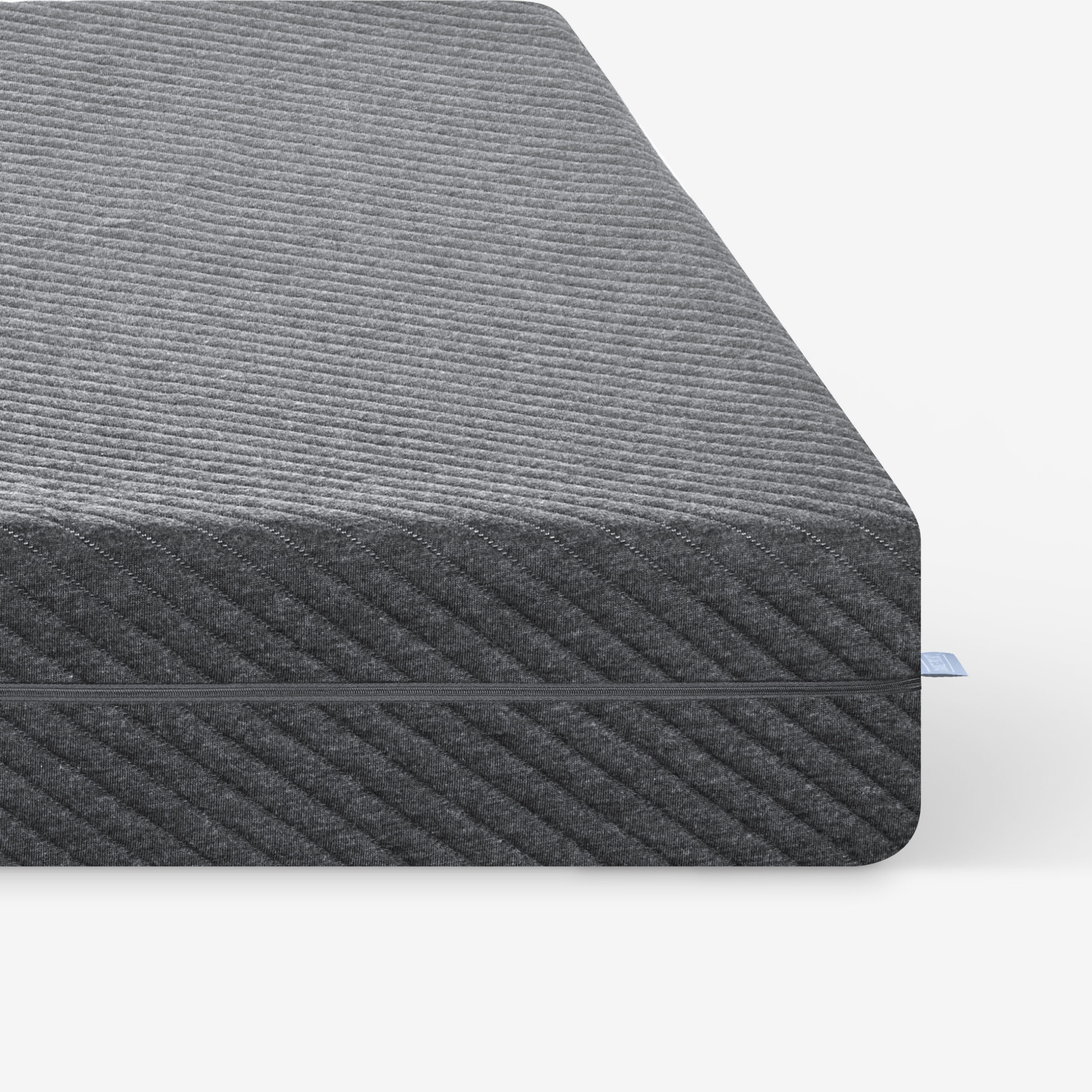Nahaufnahme der Ecke einer dunkelgrauen, strukturierten Matratze mit einem dezenten diagonalen Streifenmuster vor einem schlichten weißen Hintergrund. Ein kleines blaues Stoffetikett ist an der Matratzennaht sichtbar.