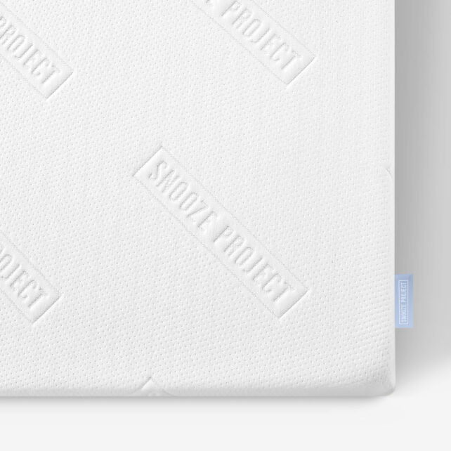 Nahaufnahme einer weißen Matratze mit strukturierter Oberfläche, auf der der Schriftzug SKOOZE PROJECT eingeprägt ist. Am rechten Rand ist ein kleines blaues Stoffetikett zu sehen.