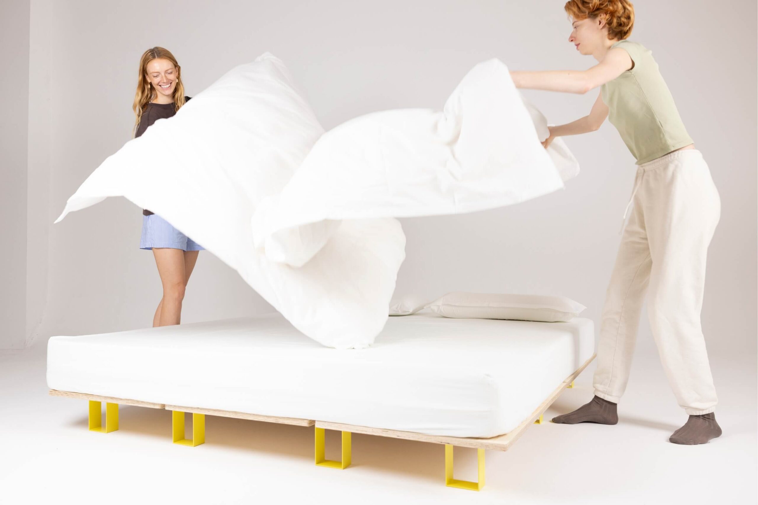 Zwei Personen machen gemeinsam ein Bett, wobei sie eine weiße Bettdecke über ein ordentlich gemachtes Bett mit einem einfachen Holzrahmen und gelben Beinen in einem hellen, minimalistischen Raum ausbreiten.