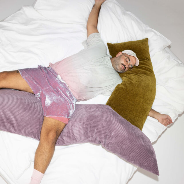 Eine Person, die ein helles Hemd, rosa Shorts und eine Mütze trägt, liegt ausgestreckt auf einem Bett mit weißen Laken und ruht sich auf großen grünen und lila Kissen aus.