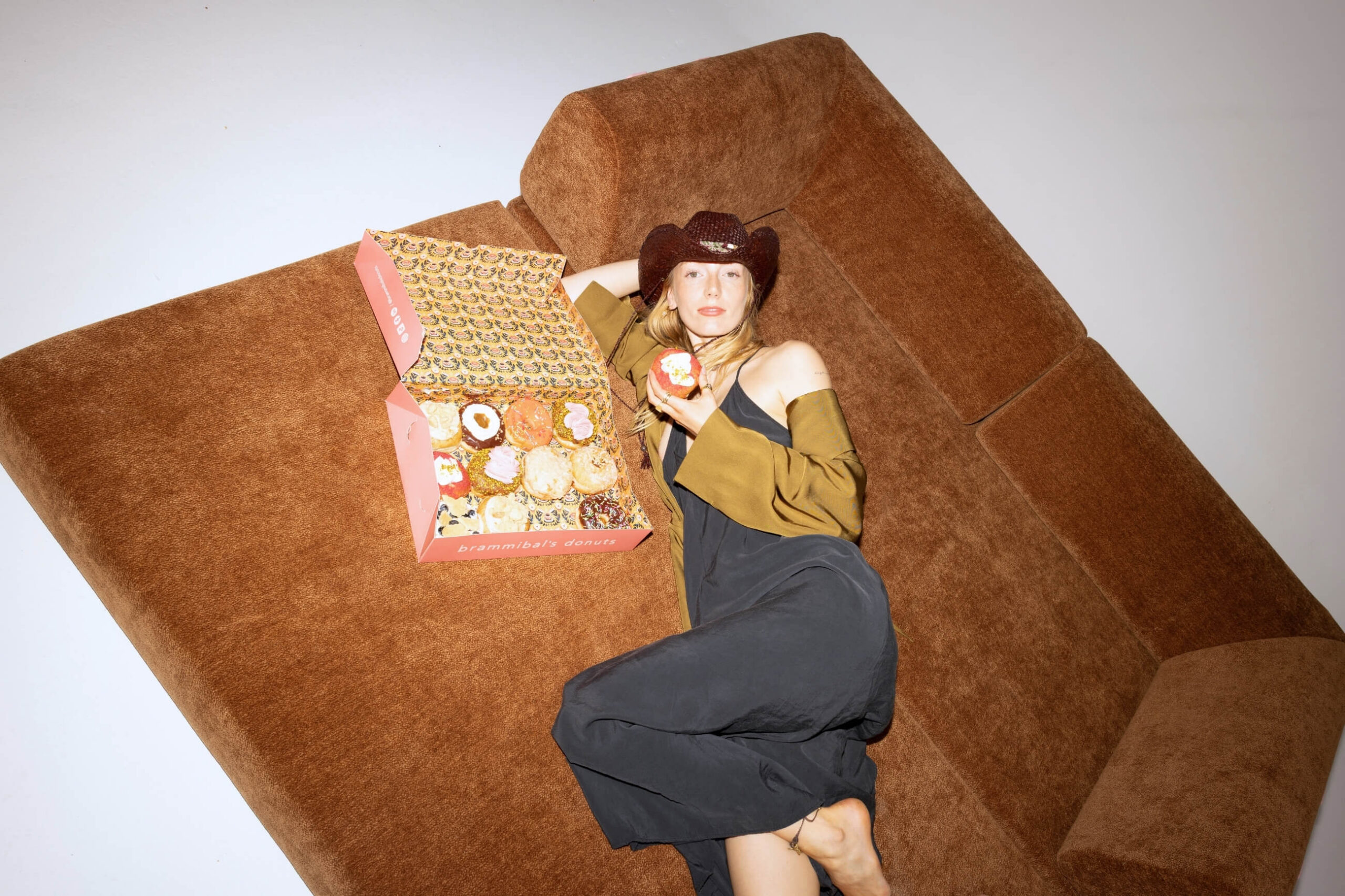 Eine Frau in einem schwarzen Kleid und einem braunen Cowboyhut liegt barfuß auf einem braunen Sofa und hält einen Donut in der Hand. Eine rosa Schachtel mit verschiedenen Donuts liegt neben ihr auf dem Sofa.